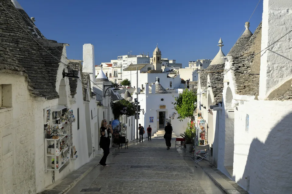 A cobbled street climbing between trulli with grey conical stone roofs in the Rione Monti at Alberobello, with souvenir stalls and visitors along it