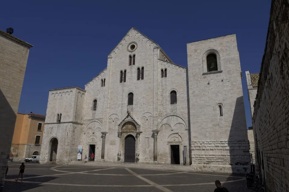 The Romanesque stone facade of the Basilica di San Nicola in Bari Vecchia, flanked by its two towers, seen from the paved square in front