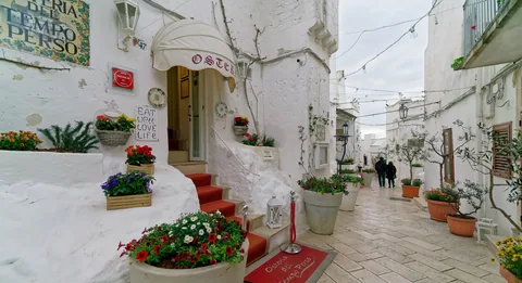 A narrow whitewashed lane in the old town of Ostuni lined with flower pots and potted olive trees, with the red-carpeted steps of a restaurant entrance on the left