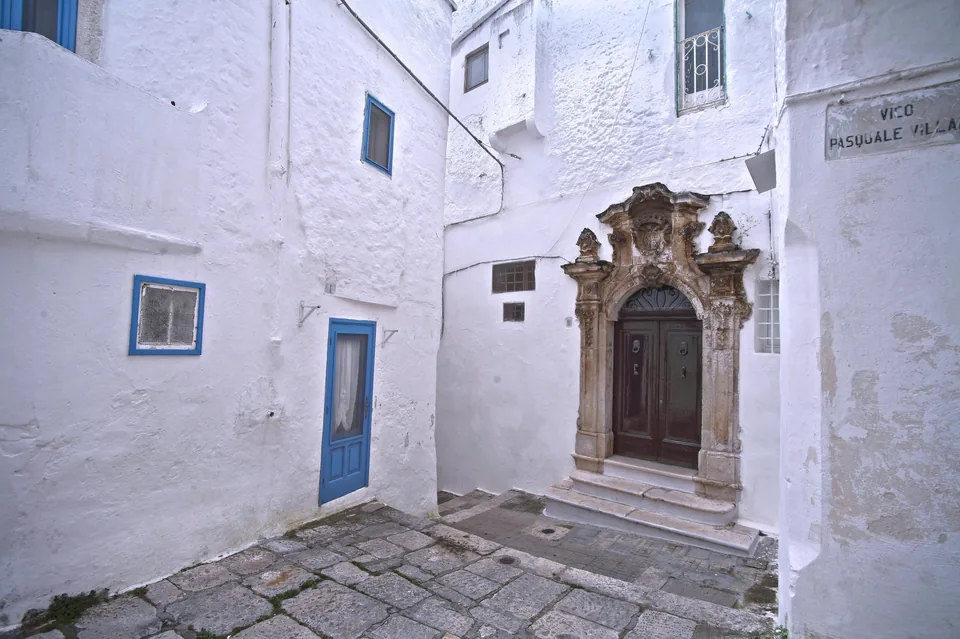 A whitewashed dead-end courtyard in the old town of Ostuni, with a carved Baroque stone doorway, blue-painted doors and windows and a flagstone floor