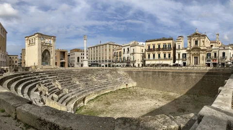 The excavated Roman amphitheatre in Piazza Sant'Oronzo, Lecce, with its tiers of stone seating, the column of Sant'Oronzo and the Sedile behind