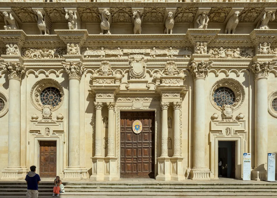 The lower half of the Baroque facade of the Basilica di Santa Croce in Lecce, with its carved portal, the two side rose windows and the figured frieze in Lecce stone