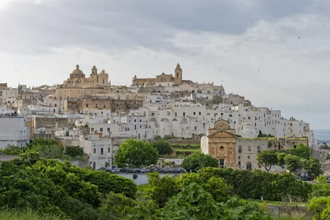 The white old town of Ostuni stacked on its hill, with the cathedral dome and a Baroque church front, seen from below across dense green woodland