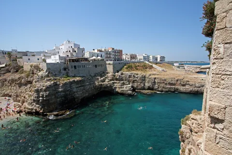 The bay of Cala Porto at Polignano a Mare crowded with swimmers on a summer day, below the limestone cliff with the white old town built along its edge