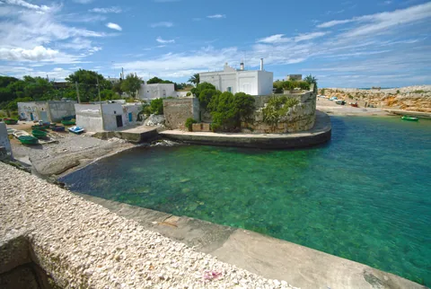 Cala Paura at Polignano a Mare, with small wooden fishing boats drawn up on the shingle, low fishermen's huts and a whitewashed house on the walled headland